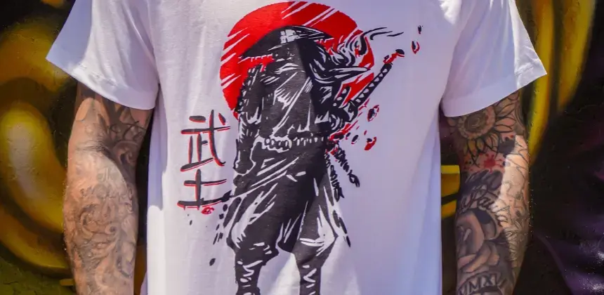 Camiseta blanca con diseño de samurái y sol rojo, usada por persona con tatuajes en los brazos