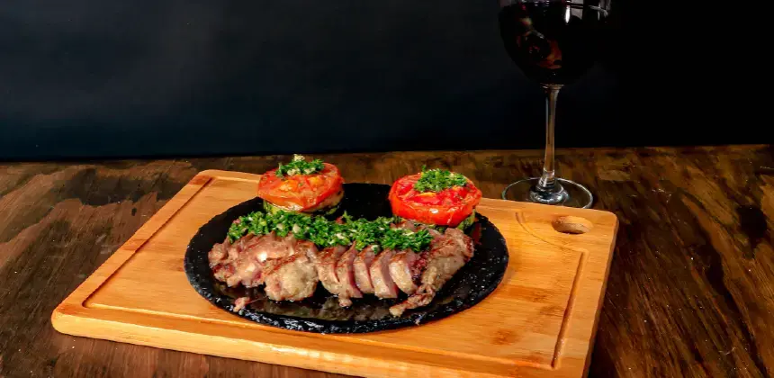 Plato de carne asada con tomates al horno y copa de vino tinto sobre tabla de madera