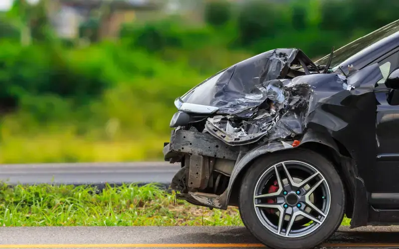 Vehículo negro con la parte frontal gravemente dañada tras un accidente, detenido al costado de una carretera con vegetación de fondo.