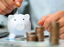 Persona insertando una moneda en una alcancía blanca con pilas de monedas en primer plano, representando el ahorro financiero.