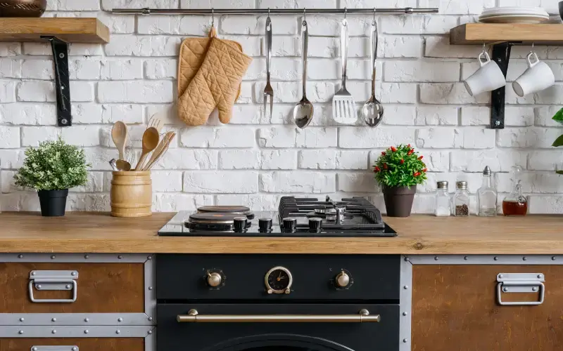 Cocina rústica con encimera de madera, utensilios colgantes, estufa y plantas decorativas frente a una pared de ladrillo blanco