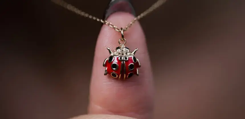 Collar dorado con dije de mariquita roja sostenido frente a un dedo en primer plano.