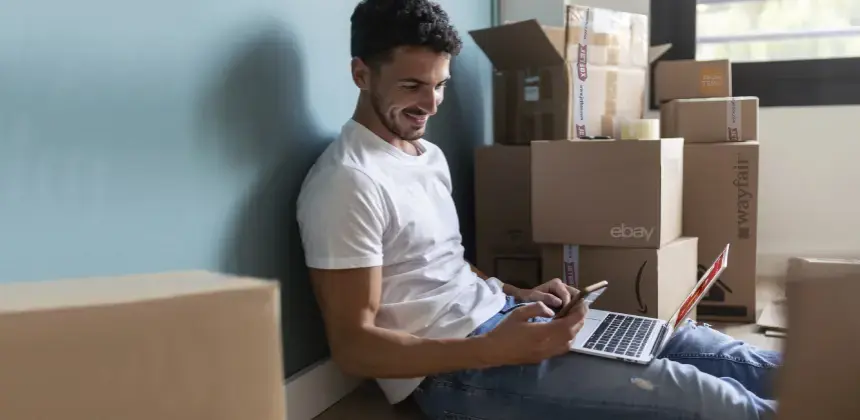 Hombre sentado en el suelo entre cajas de envío usando una laptop y una tarjeta de crédito para compras en línea