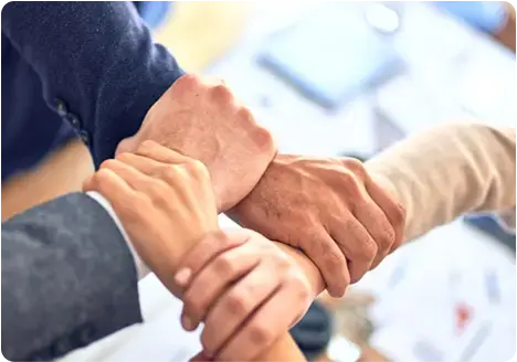 Cuatro personas entrelazando sus brazos en señal de trabajo en equipo y unión, con fondo desenfocado de oficina.