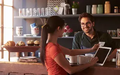 Mujer realizando un pedido digital en una cafetería, mientras el barista sonríe detrás del mostrador con productos horneados al frente.