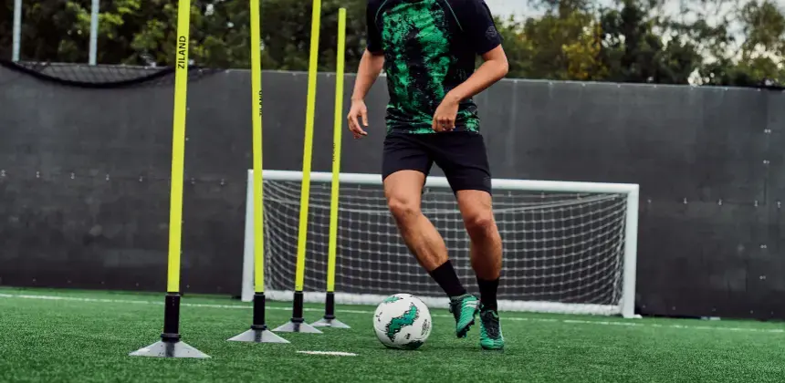 Jugador de fútbol entrenando con conos y pelota en una cancha de césped sintético