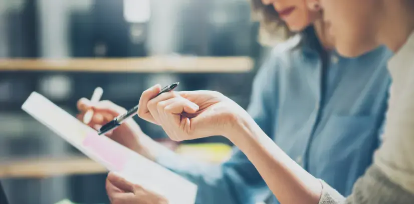 Dos personas revisando y señalando información en una hoja con bolígrafos en un entorno de oficina.