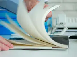 Persona hojeando un cuaderno en una oficina con teléfono y carpetas al fondo.