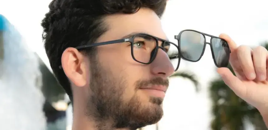 Hombre con barba sosteniendo unas gafas de sol frente a su rostro mientras usa anteojos ópticos