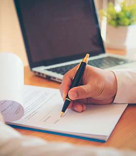 Persona firmando un documento sobre un escritorio con una computadora portátil al fondo.