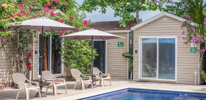 Área de piscina con sillas, mesas y sombrillas frente a cabañas rodeadas de plantas.