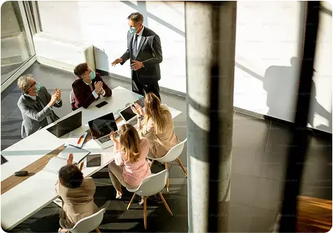 Reunión empresarial con personas usando mascarillas sentadas alrededor de una mesa mientras un hombre de pie realiza una presentación.