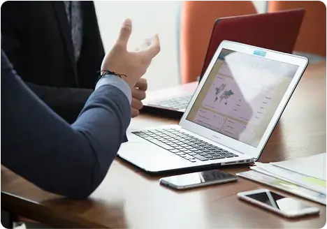 Persona explicando datos en una laptop con gráficos y mapas en pantalla, acompañada de celulares y documentos sobre una mesa de trabajo.