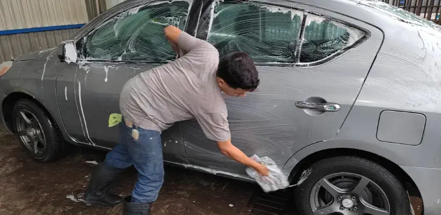 Persona lavando un automóvil gris con esponja y jabón en un autolavado