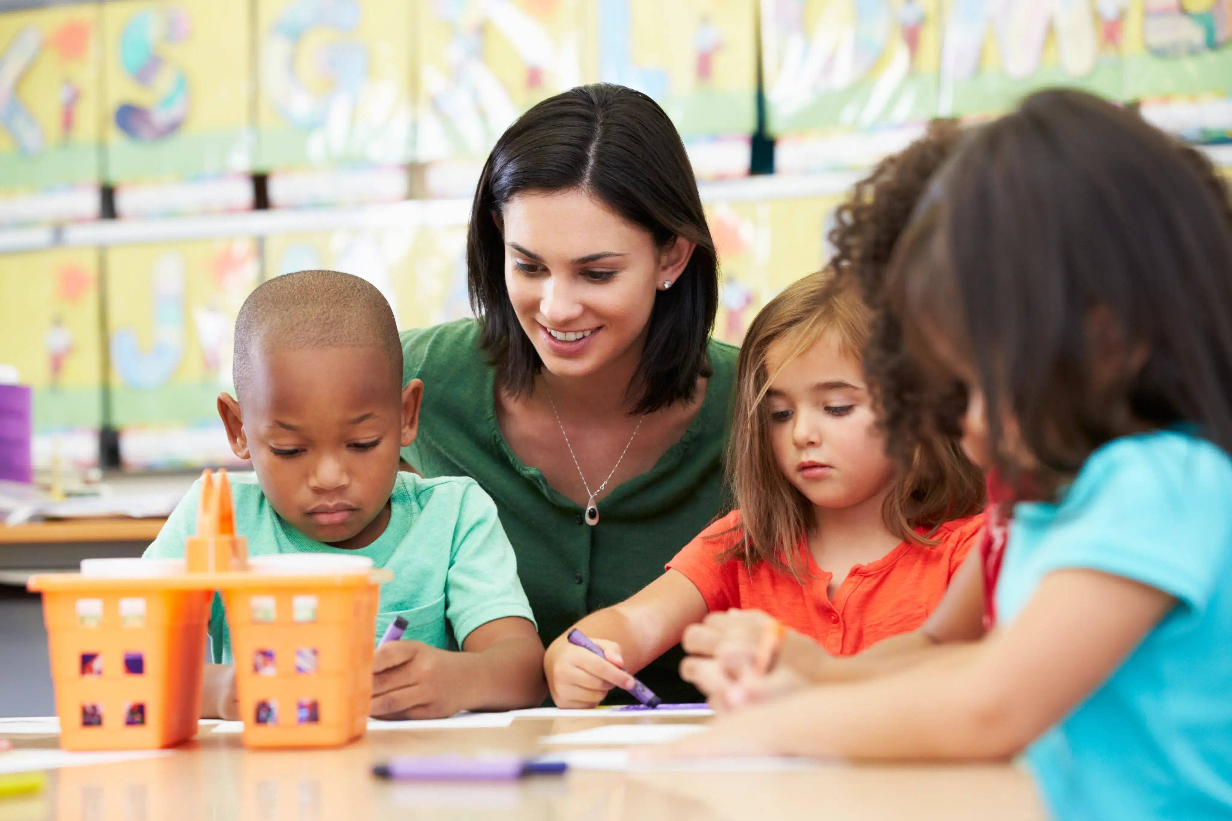 Maestra observa y guía a varios niños pequeños mientras dibujan en una mesa de aula.