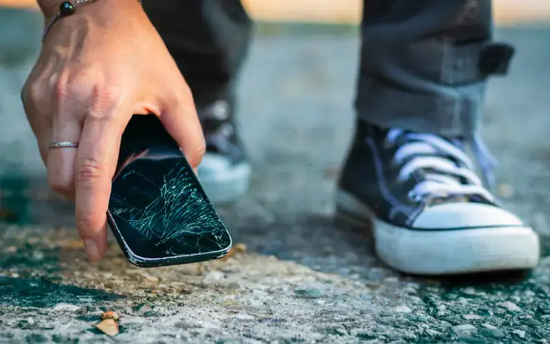 Persona recogiendo un celular con la pantalla rota del suelo, con enfoque en el daño y los zapatos tipo converse.