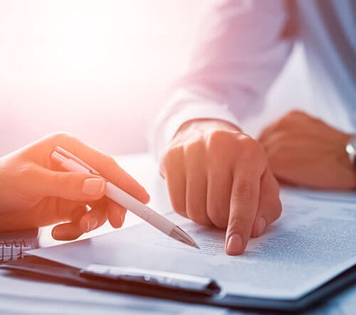 Persona señalando un documento con un bolígrafo sobre una mesa iluminada.