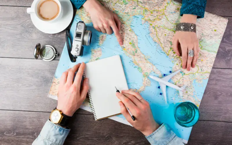 Manos planificando unas vacaciones sobre un mapa, con accesorios de viaje como cámara, avión de juguete y brújula sobre la mesa