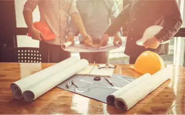 Mesa de trabajo con planos y equipo de seguridad, mientras profesionales de la construcción revisan un proyecto.