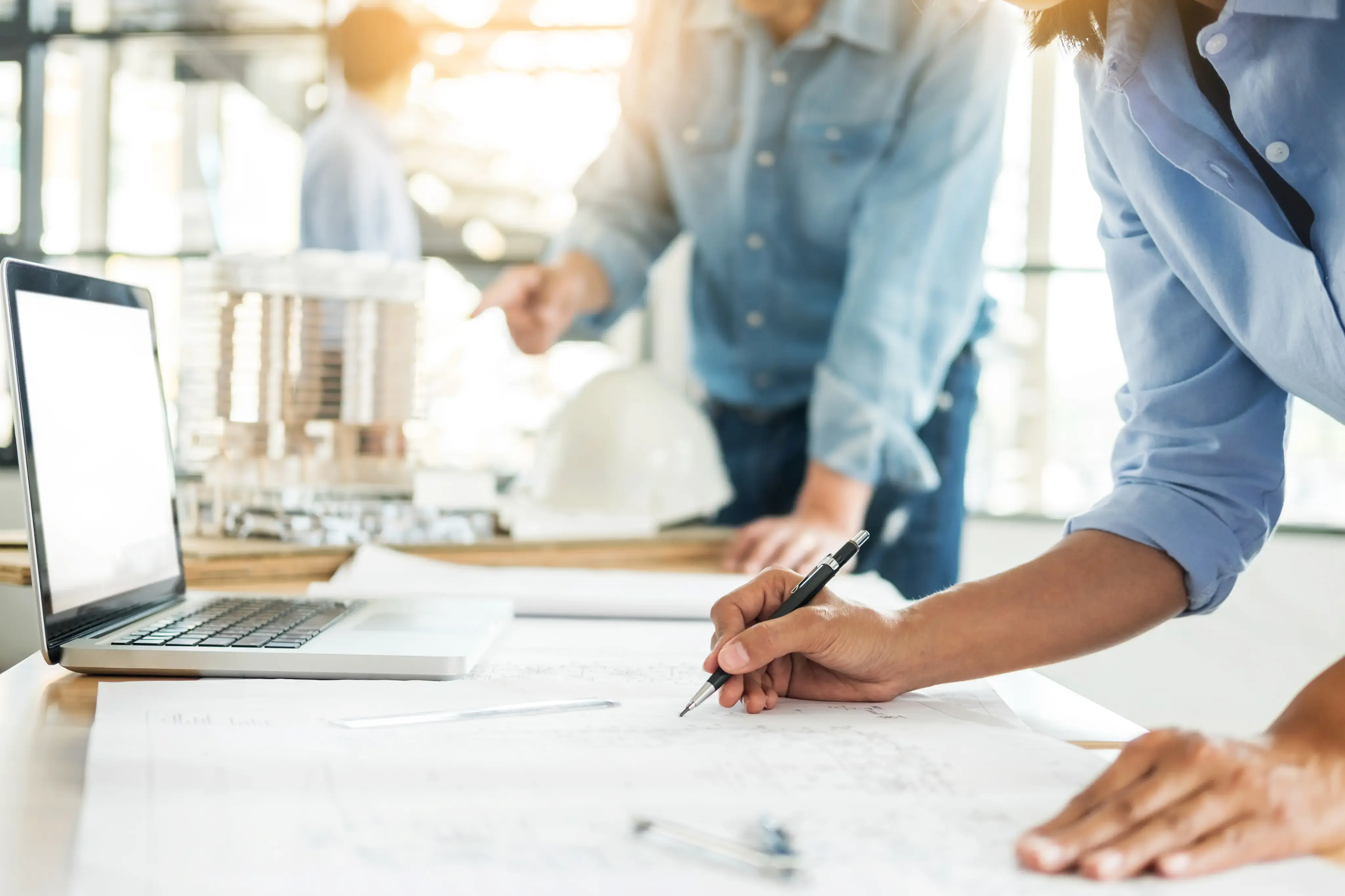Persona tomando notas sobre planos arquitectónicos en una mesa con laptop y maqueta.