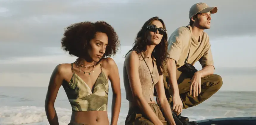 Tres personas posando con ropa casual en tonos tierra frente al mar durante el atardecer