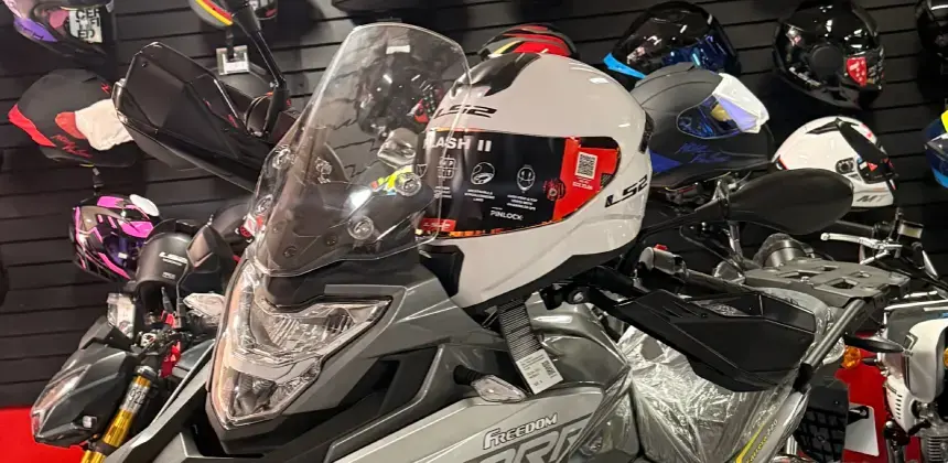 Motocicleta gris en exhibición con un casco blanco en el manillar y varios cascos adicionales colgados en la pared de fondo.