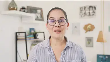 Mujer con gafas y camisa de rayas frente a una pared decorada, hablando directamente a la cámara en un entorno tipo oficina o estudio.