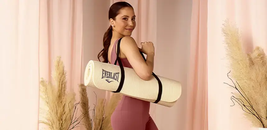 Mujer con atuendo deportivo color rosa cargando un tapete de ejercicio Everlast en un entorno interior