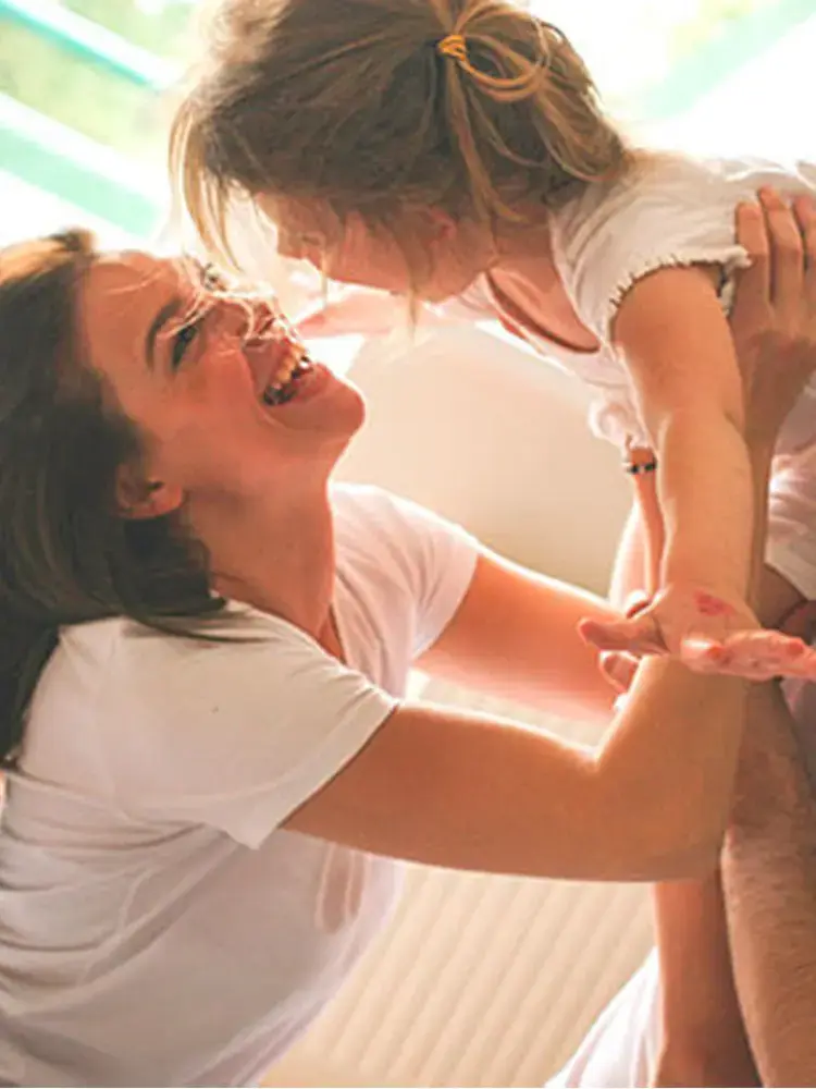 Madre levantando con alegría a su hija pequeña en el aire dentro de casa, compartiendo un momento de ternura y felicidad familiar.