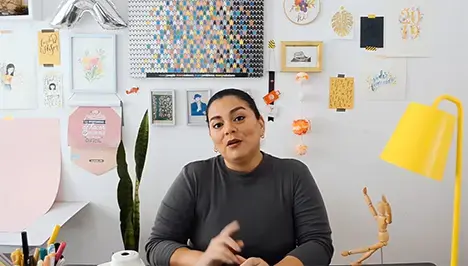 Mujer sentada en un escritorio colorido, con decoraciones artísticas en la pared detrás de ella, hablando a la cámara.