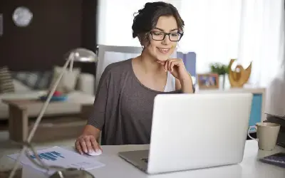 Mujer con gafas sentada frente a una computadora portátil, usando un mouse sobre el escritorio, con papeles y una taza al lado.