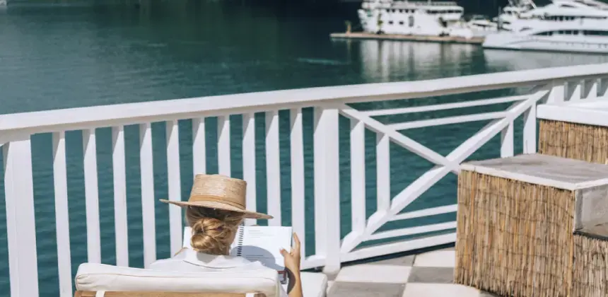 Mujer con sombrero leyendo un libro frente al mar en una terraza con vista a yates