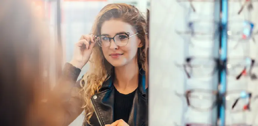 Mujer probándose gafas frente a un espejo en una óptica con exhibición de monturas al fondo