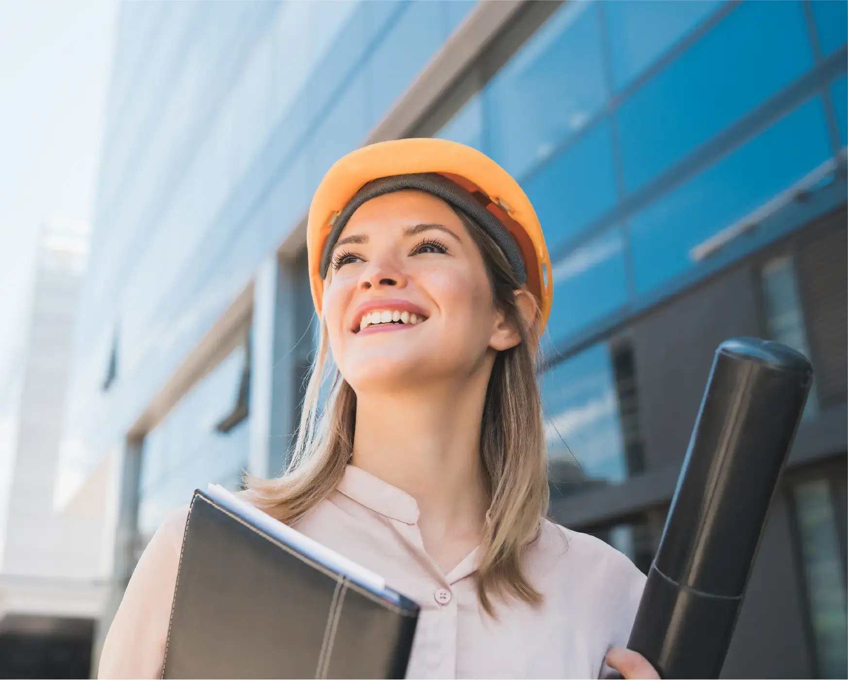 Mujer joven con casco de seguridad sonriendo mientras sostiene documentos y planos frente a un edificio moderno.