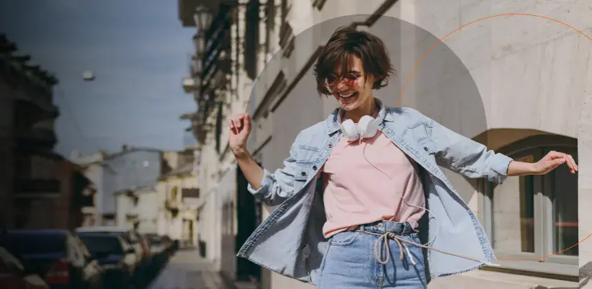 Mujer joven sonriente caminando por la ciudad con audífonos y ropa casual de mezclilla
