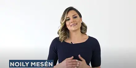 Noily Mesén hablando frente a una pared blanca, vistiendo blusa oscura y con micrófono de solapa.