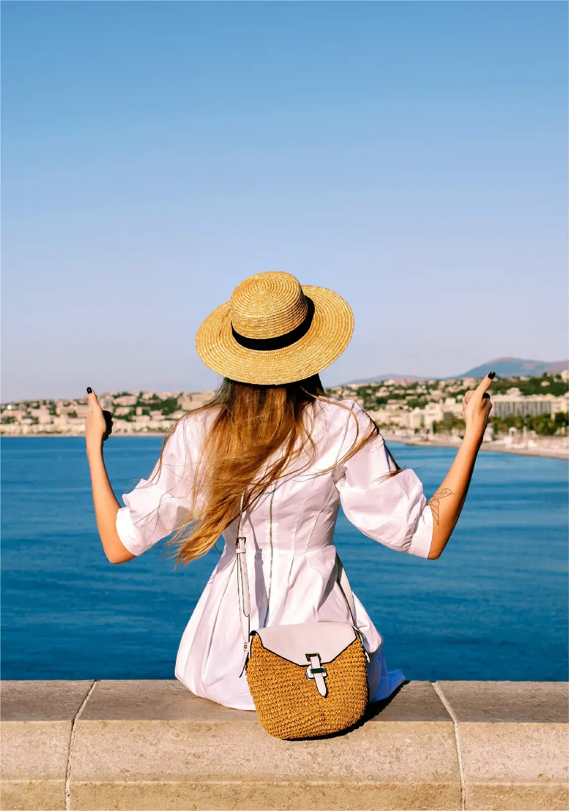 Mujer de espaldas sentada frente al mar con sombrero de paja, levantando los brazos con un gesto de libertad mientras contempla el paisaje.