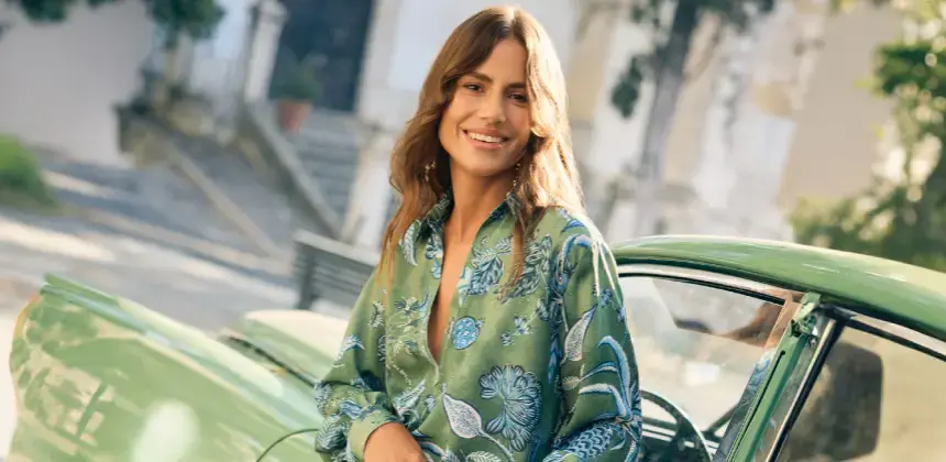 Mujer sonriente con blusa verde de estampado floral posando junto a un automóvil clásico