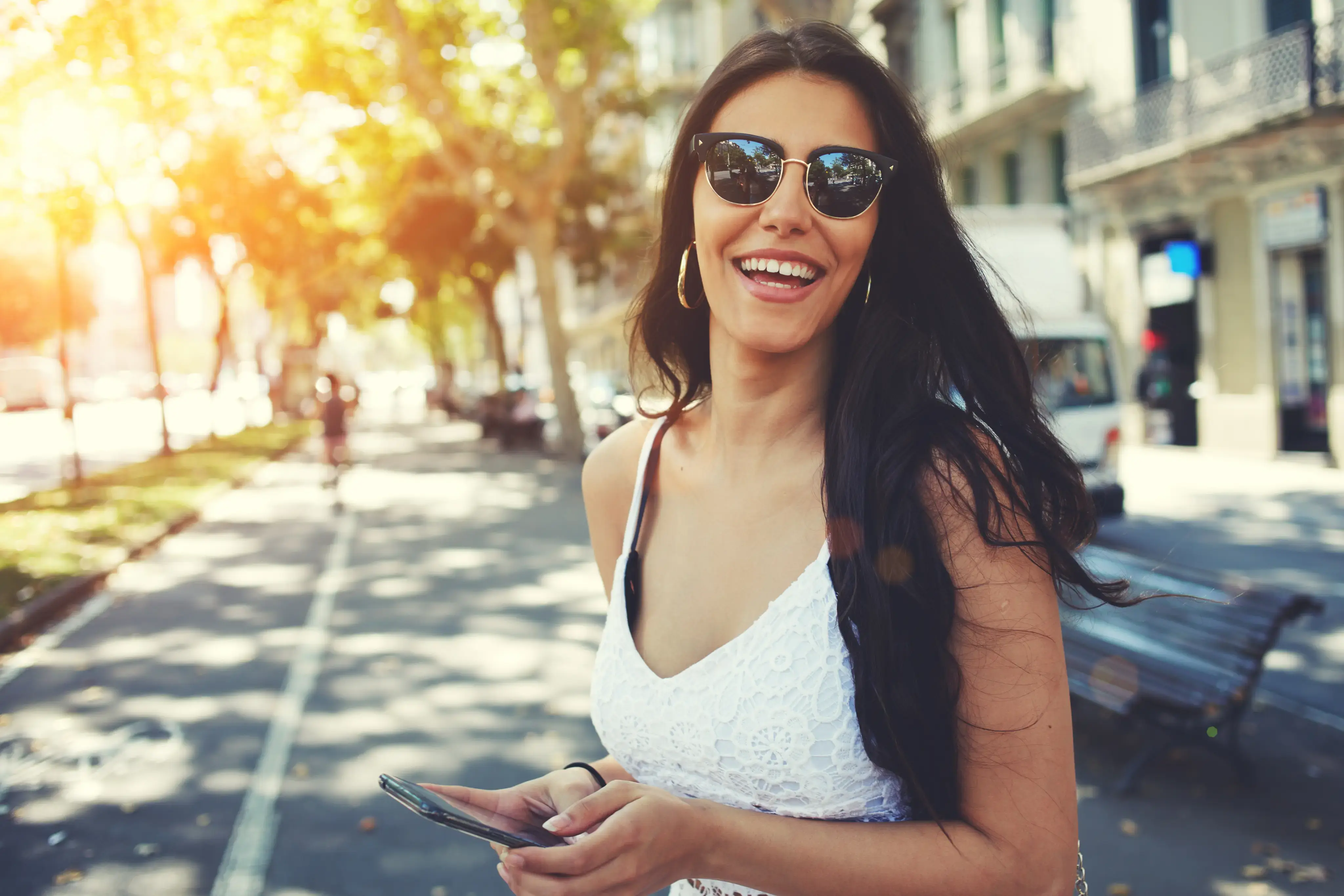 Mujer joven con gafas de sol sonriendo mientras sostiene un teléfono en una calle soleada.