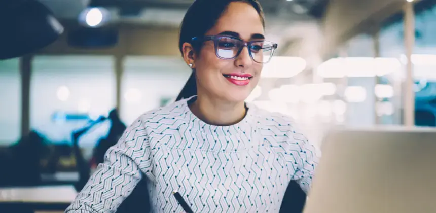 Mujer con gafas trabajando frente a una computadora portátil en una oficina iluminada