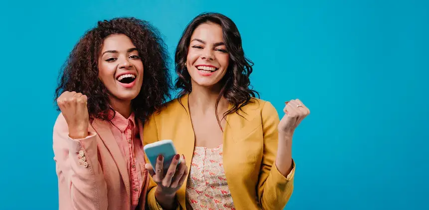 Dos mujeres sonrientes celebrando mientras una sostiene un teléfono móvil sobre fondo azul