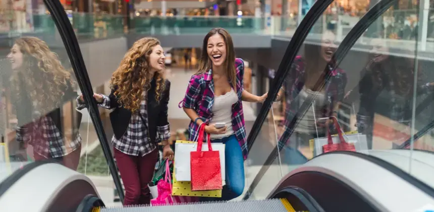 Dos mujeres sonrientes subiendo por una escalera eléctrica con bolsas de compras en un centro comercial