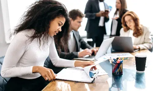 Mujer revisando documentos en una sala de reuniones, acompañada de compañeros trabajando con computadoras portátiles
