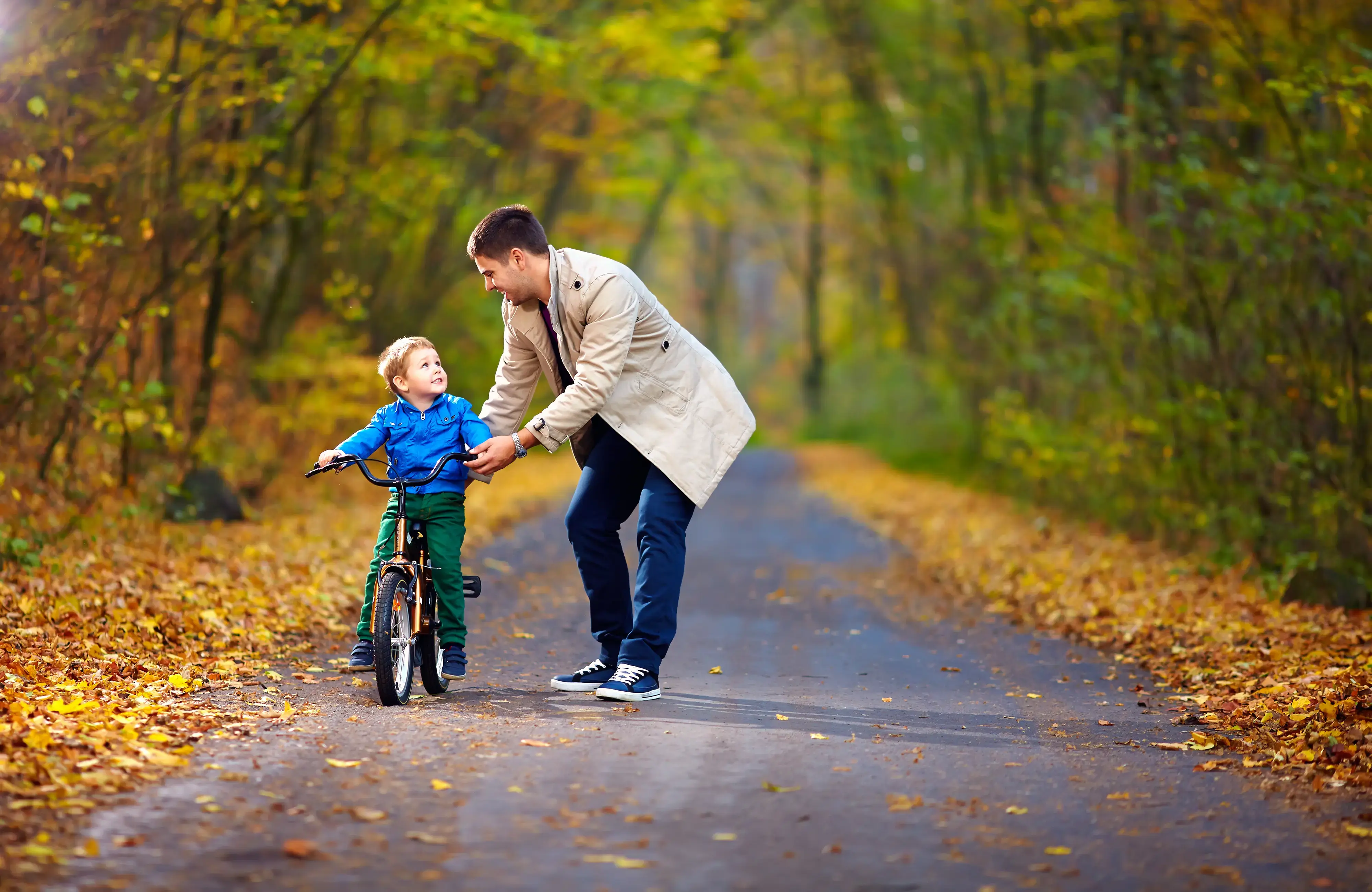 Hombre ayudando a un niño a montar una bicicleta en un camino con hojas otoñales.