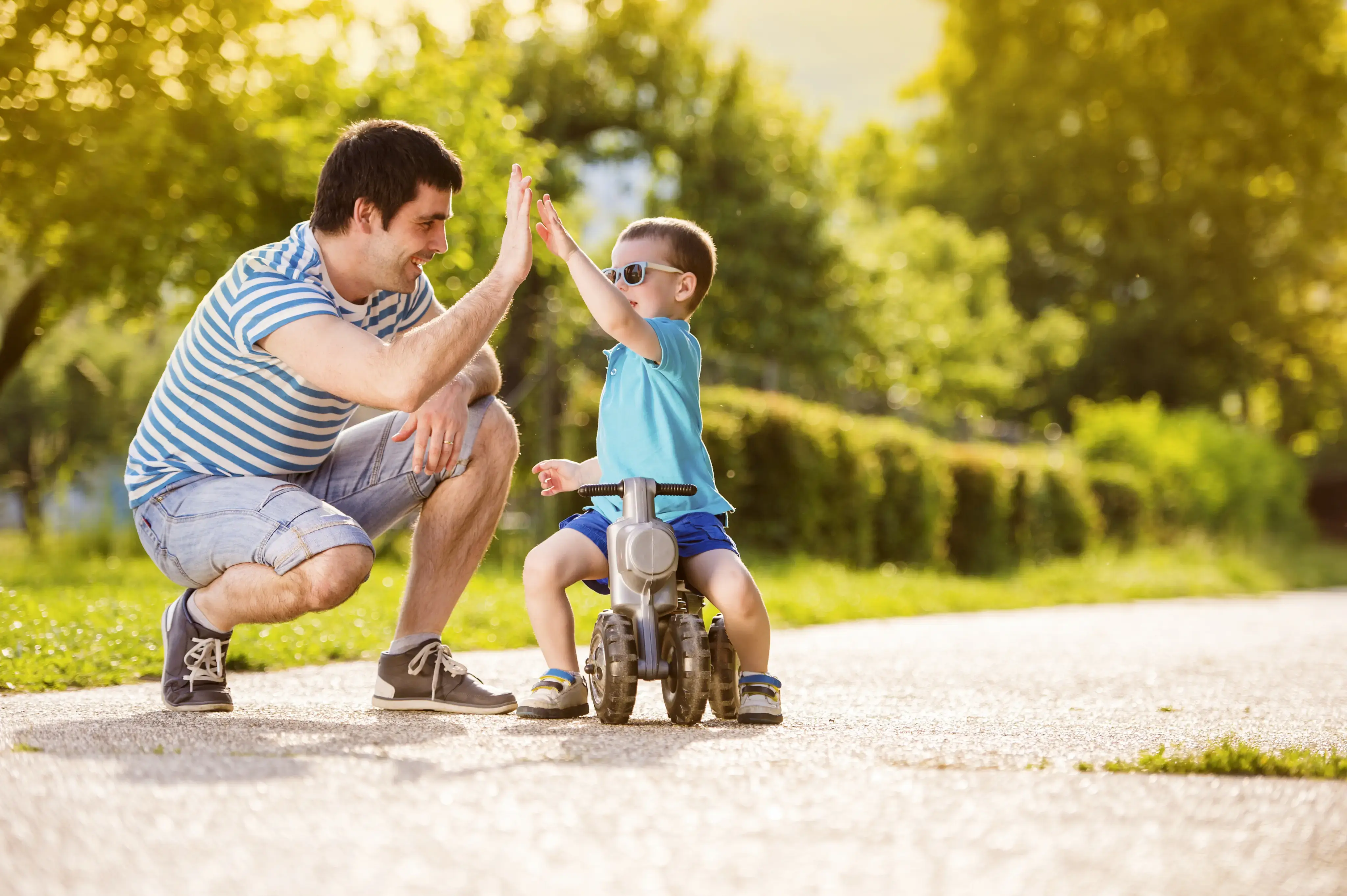 Padre e hijo chocando las manos mientras el niño está sentado en un juguete con ruedas al aire libre.