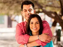 Pareja joven abrazándose y sonriendo al aire libre con árboles desenfocados al fondo.
