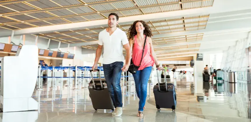 Pareja caminando por el aeropuerto con maletas de viaje de mano, sonrientes y tomados de la mano