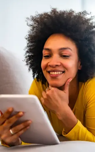 Mujer sonriente recostada en un sofá, usando una tableta digital con actitud relajada y positiva.