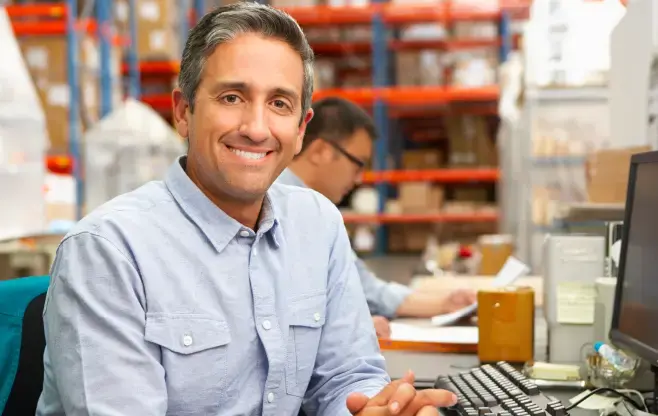 Hombre adulto sonriendo frente a una computadora en un almacén, con estantes llenos al fondo y otro trabajador revisando documentos.