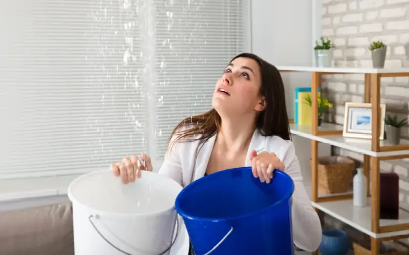Mujer dentro de una casa mirando al techo con preocupación mientras sostiene dos baldes para contener una filtración de agua.